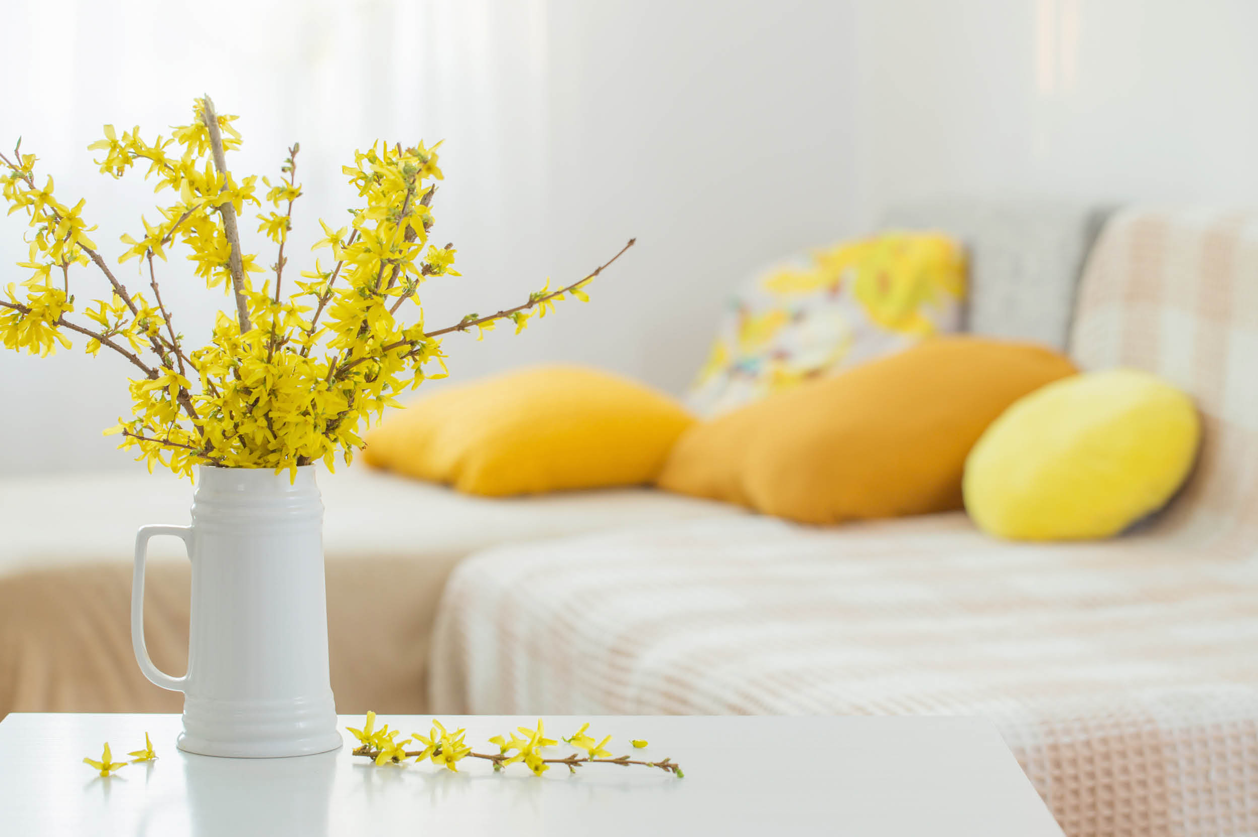 A white vase with yellow flowers sits on a table in front of a couch with yellow pillows. AI generated content