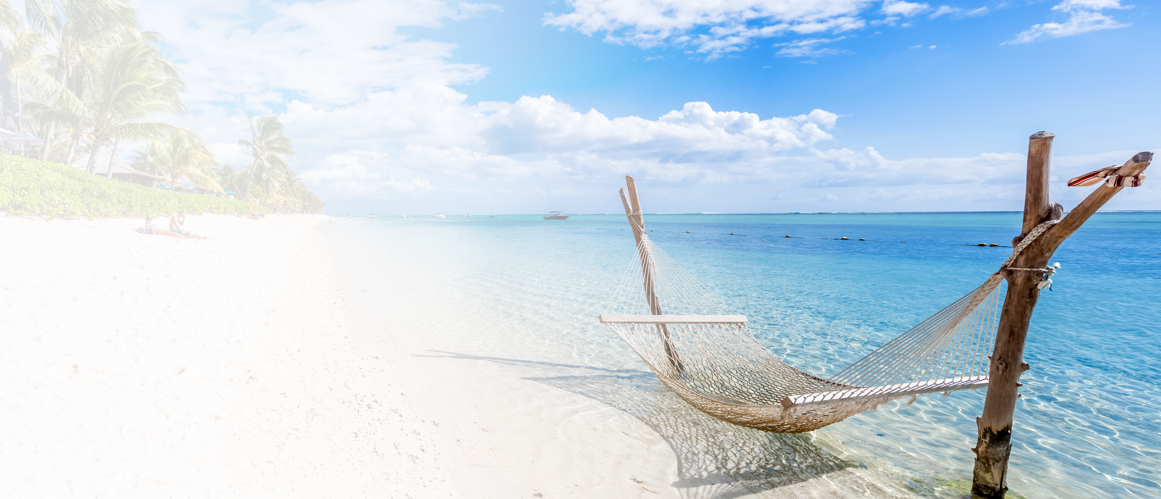 A hammock is suspended over clear blue water near a sandy beach with palm trees and people relaxing in the background under a partly cloudy sky. Description generated by AI